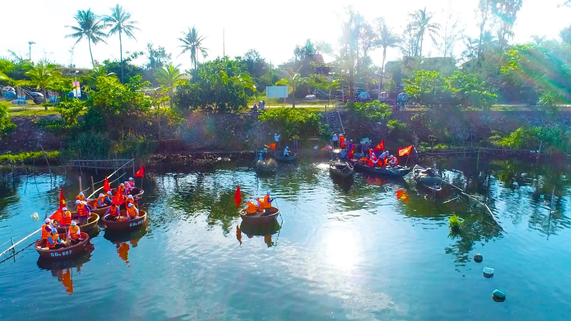 Nhiều thúng chai chở khách mặc áo phao cam, cắm cờ đỏ, chèo trên mặt nước giữa rặng dừa và bờ kè đá