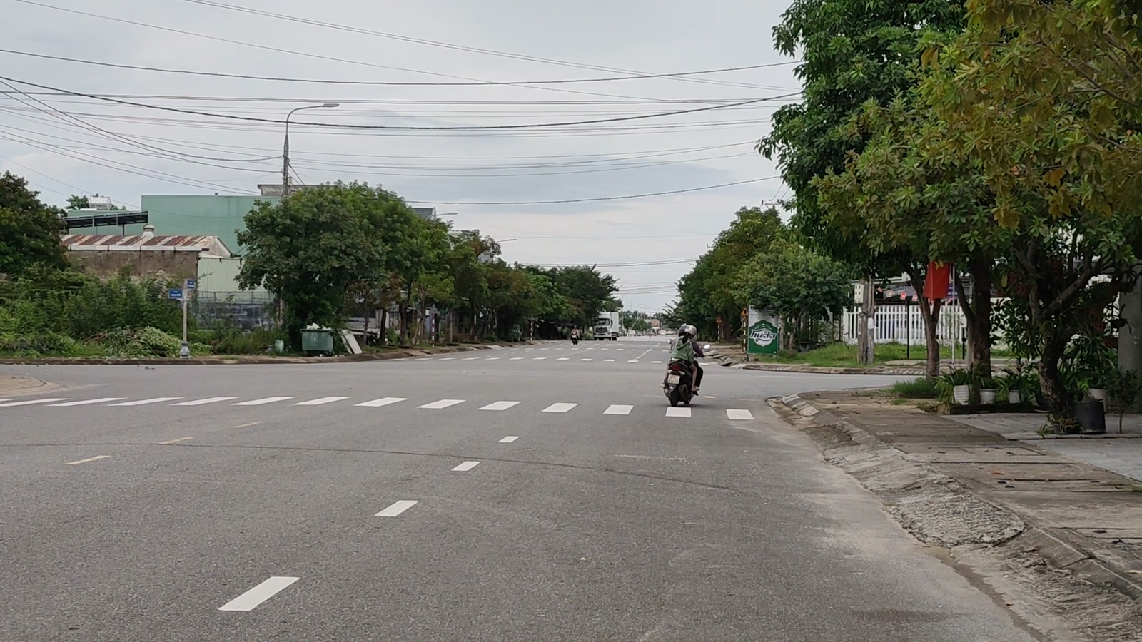 Ngã tư rộng có vạch qua đường, một người chạy xe máy băng qua, bên phải là vỉa hè rộng và hàng cây xanh
