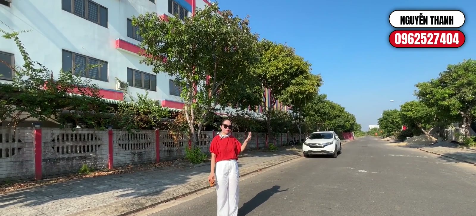 Nguyễn Thanh đứng trên đường nội khu rộng trong khu dân cư An Hà Quảng Phú, một ô tô đỗ bên đường và hàng cây xanh hai bên