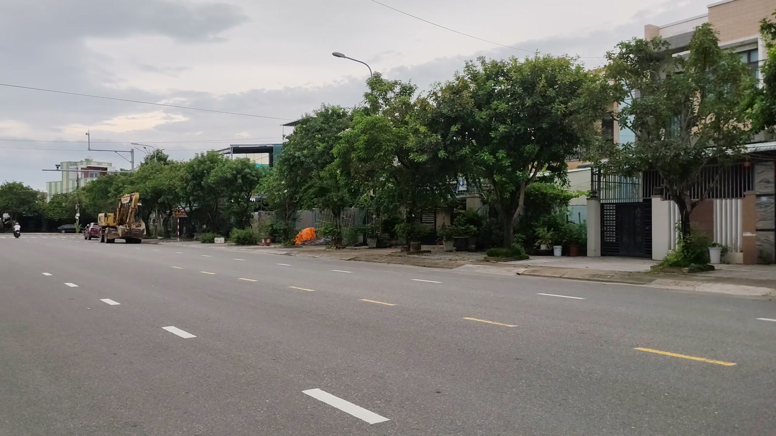 Dãy nhà phố và hàng cây dọc một con đường lớn nhiều làn xe ở Tam Kỳ, có xe công trình đậu phía xa