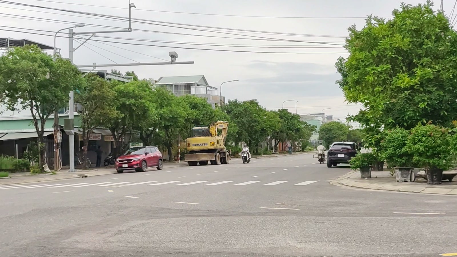 Ngã tư rộng trên đường Lê Quý Đôn Tam Kỳ, xe máy chạy qua, một bên là dãy nhà và xe công trình đậu ven đường