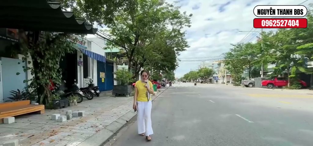 Nguyễn Thanh đứng trên mặt tiền đường Nguyễn Văn Trỗi Tam Kỳ, dãy nhà có mái hiên bên trái và lòng đường rộng