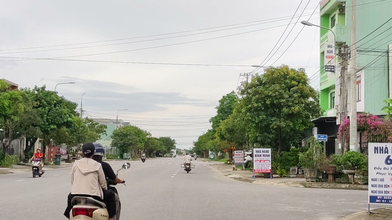 Dãy nhà nghỉ và cửa hàng với bảng hiệu dọc vỉa hè đường Lê Quý Đôn Tam Kỳ, hai người đi xe máy chạy phía trước