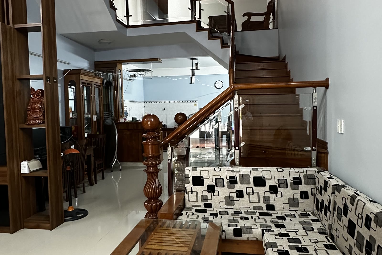 Phòng khách tầng trệt với bộ bàn ghế gỗ, kệ ti vi và cầu thang gỗ lan can kính dẫn lên tầng hai