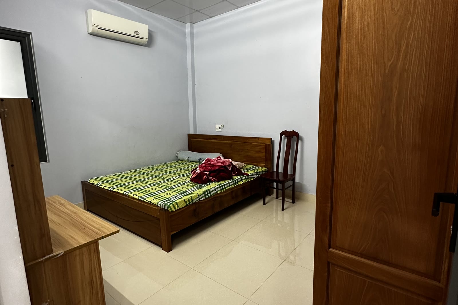 Một phòng ngủ trong nhà với giường gỗ, máy lạnh treo tường và tủ áo gỗ, nền gạch sáng