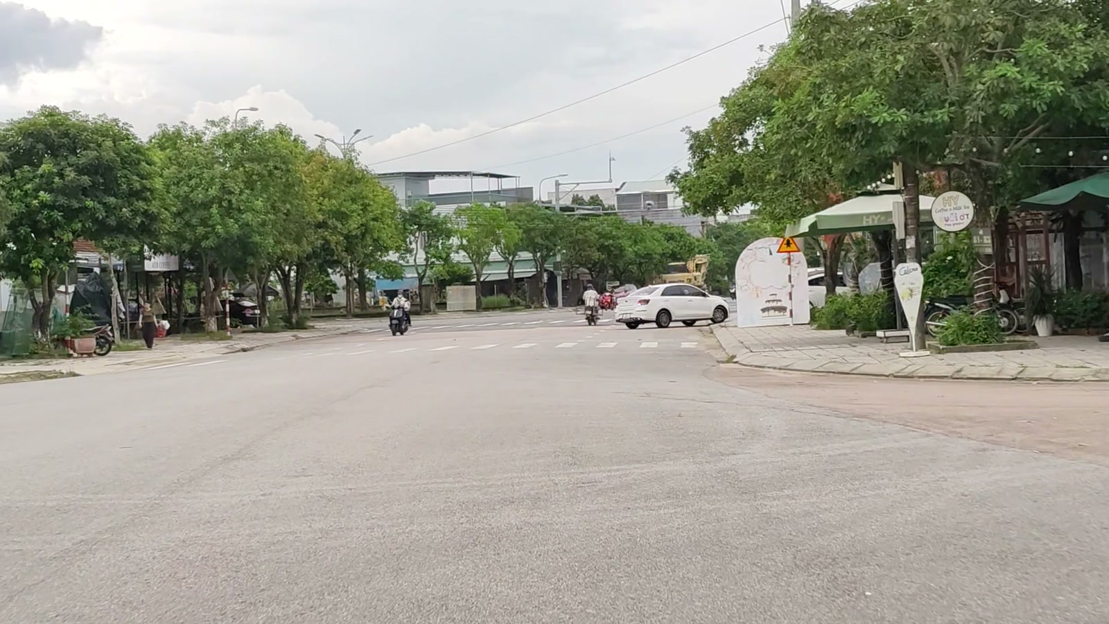 Góc ngã tư có quán cà phê che dù xanh, xe máy và ô tô chạy qua trên con đường rộng ở Tam Kỳ