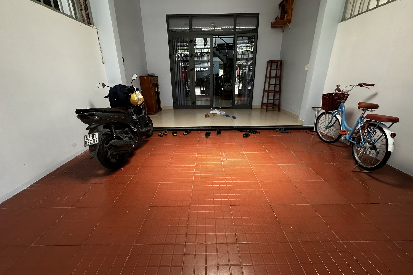 Sân để xe trong nhà lát gạch đỏ, có xe máy và xe đạp dựng bên trong, phía trước là cửa kính lớn nhìn ra đường