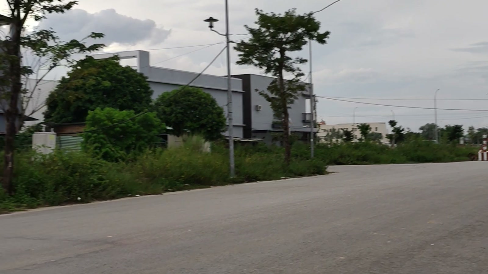 Đoạn đường rộng hướng về phía khu đất đang phát triển ở Tam Kỳ, một bên là tòa nhà có hàng rào, một bên là bãi cỏ và cây xanh