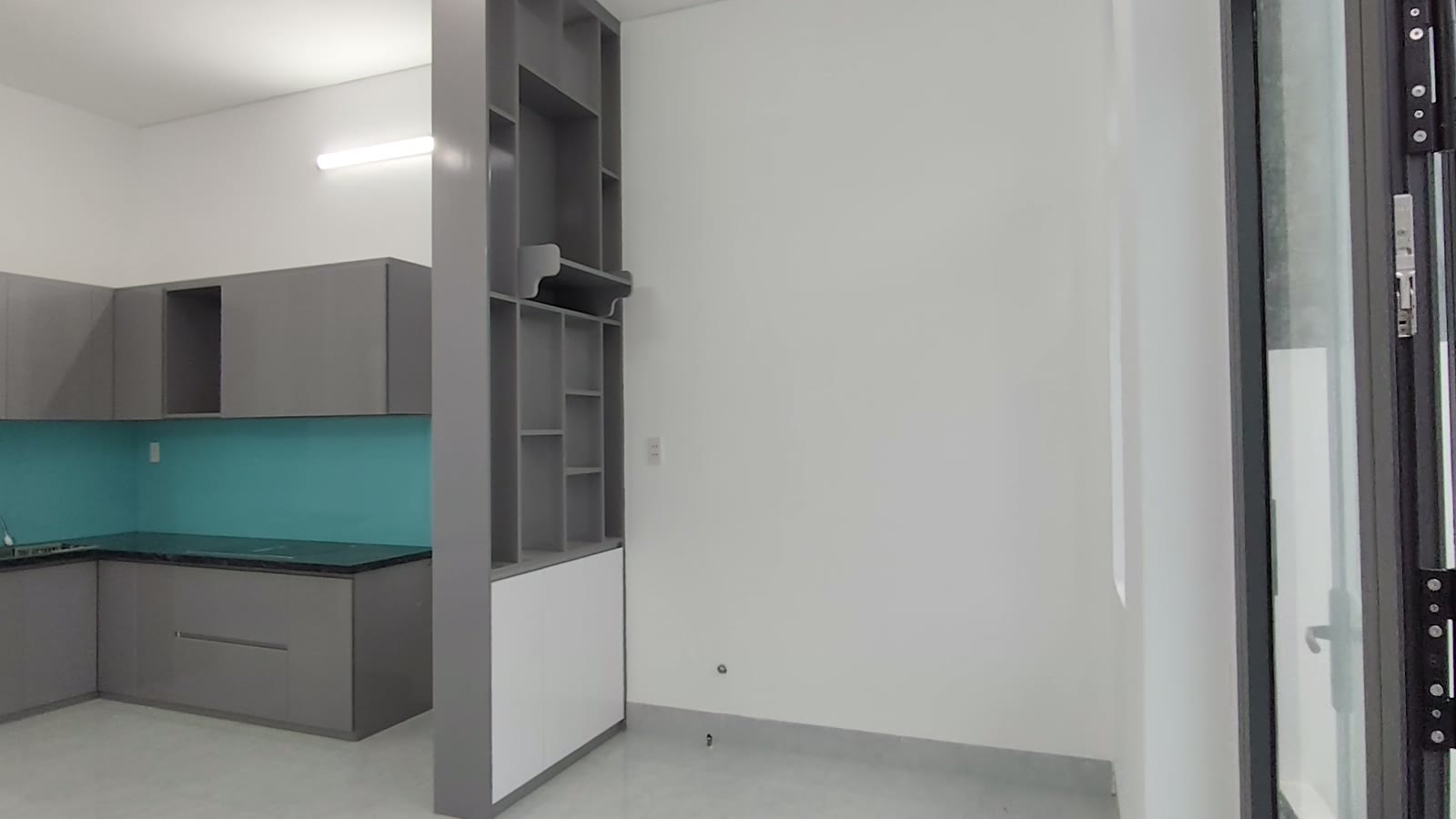 Góc bếp chữ L tủ xám ốp kính xanh và khung kệ trang trí đứng ngăn giữa bếp với phòng khách, nhà kiệt Trần Cao Vân Tam Kỳ
