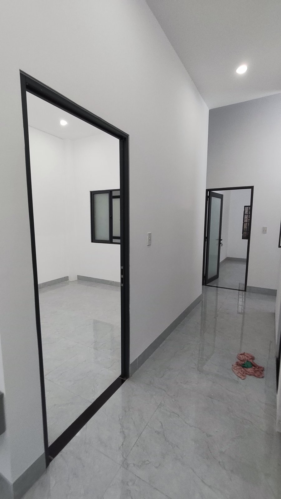 Hành lang nền gạch sáng dẫn vào các phòng ngủ, cửa khung nhôm đen mở vào phòng có cửa sổ, nhà kiệt Trần Cao Vân Tam Kỳ