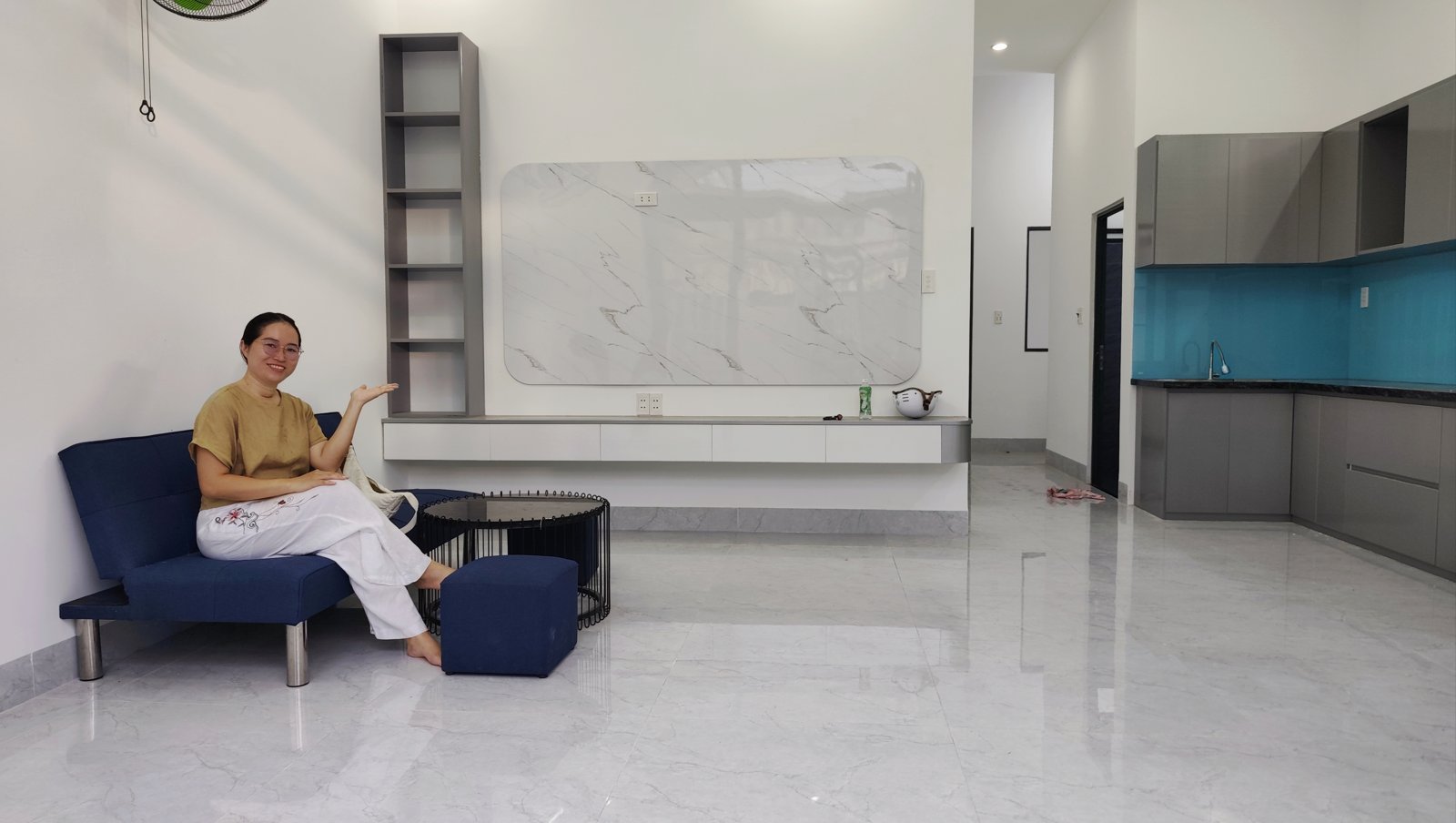 Nguyễn Thanh ngồi trên sofa xanh trong phòng khách nhà kiệt Trần Cao Vân Tam Kỳ, tay chỉ về phía bếp tủ xám ốp kính xanh ở góc phải