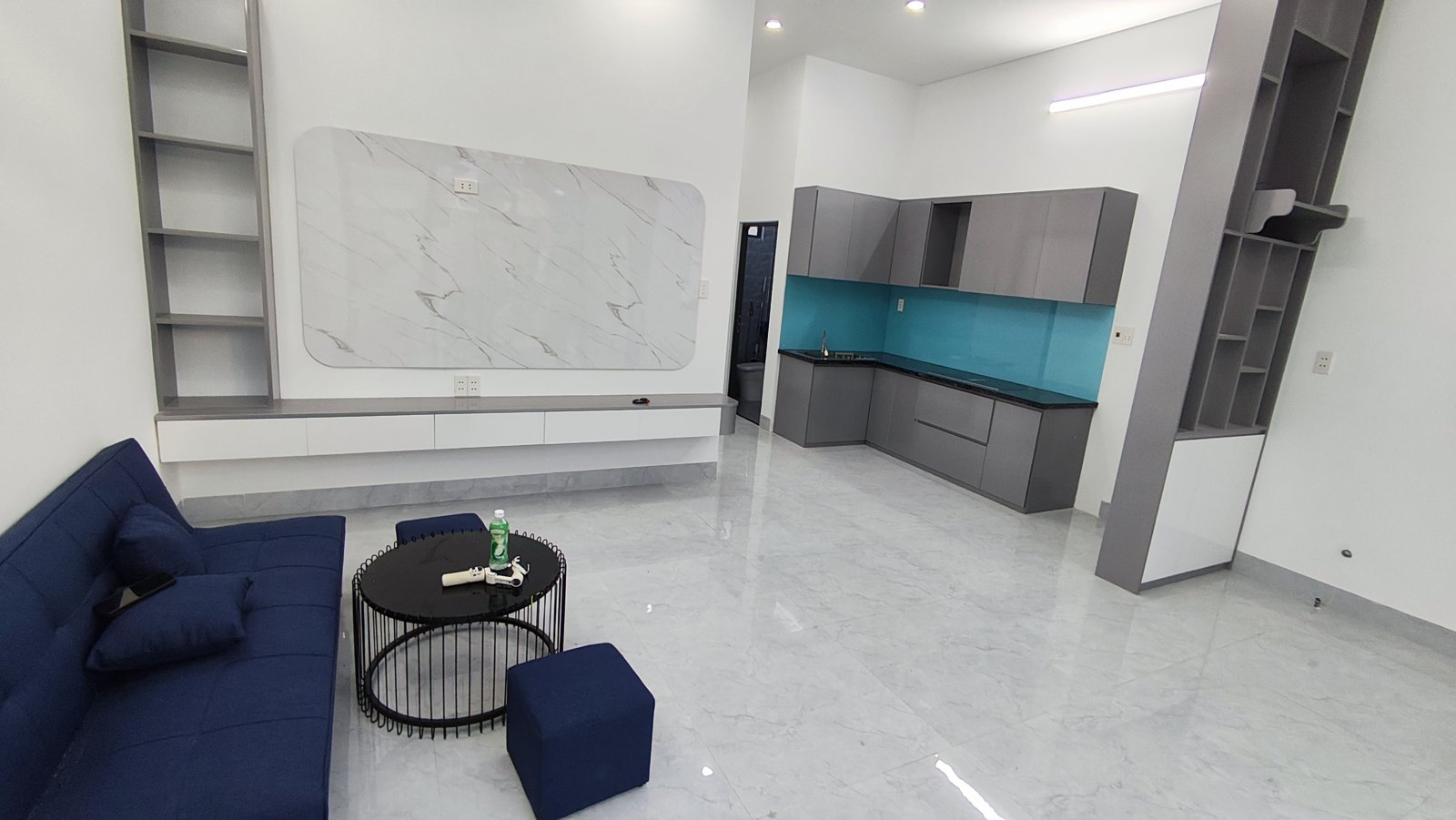 Phòng khách liên thông bếp nhà kiệt Trần Cao Vân Tam Kỳ, sofa xanh đậm, kệ tivi treo tường vân đá, bếp tủ xám ốp kính xanh phía sau