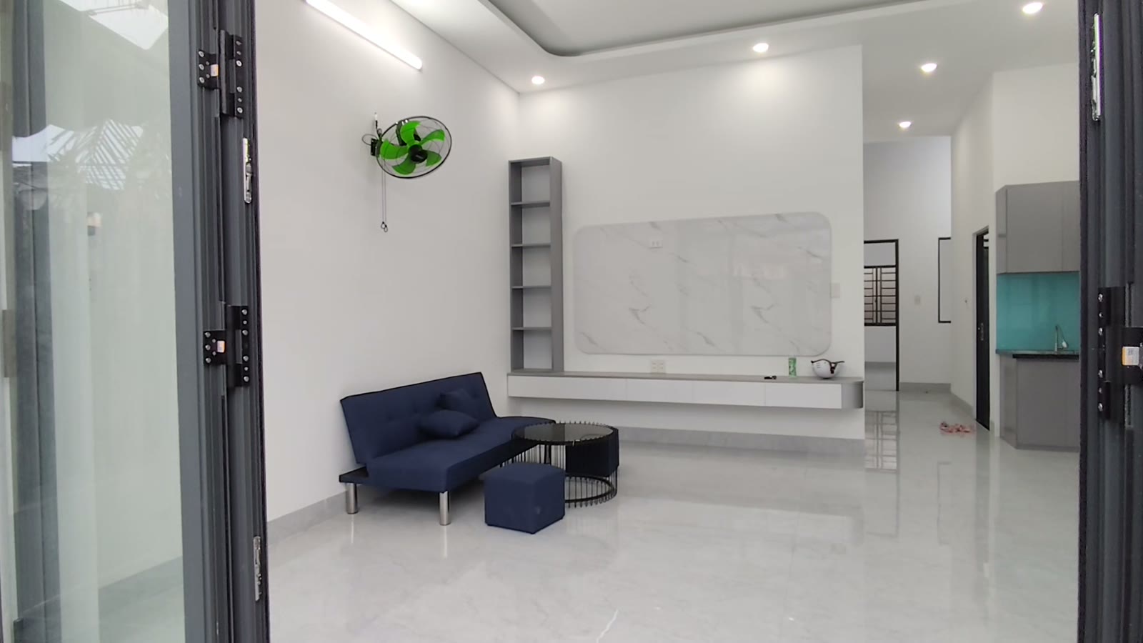 Phòng khách nhà ngang 6m ở kiệt Trần Cao Vân Tam Kỳ, sofa xanh đậm, kệ tivi vân đá treo tường, quạt treo tường, phía sau là bếp ốp kính xanh