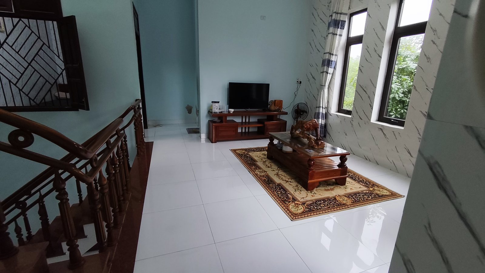 Phòng sinh hoạt chung tầng 2 có ba khung cửa sổ dài nhìn ra cây xanh, tường ốp đá vân trắng, nhà đối diện công viên Tam Kỳ