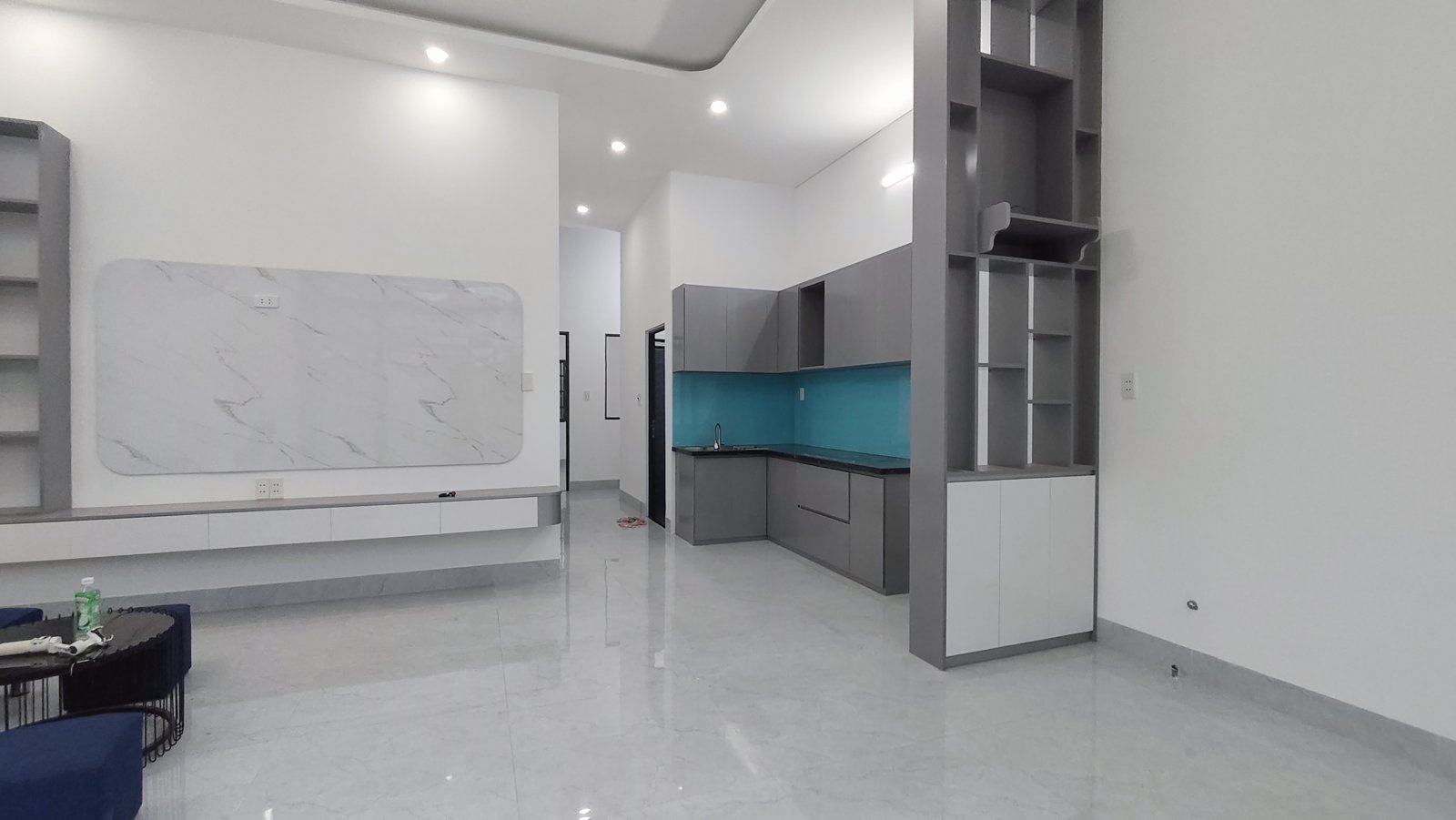 Sofa xanh, bàn tròn mặt kính đen, kệ tivi vân đá và kệ trang trí xám, bếp chữ L tủ xám ốp kính xanh ở góc phải, nhà ngang 6m Tam Kỳ