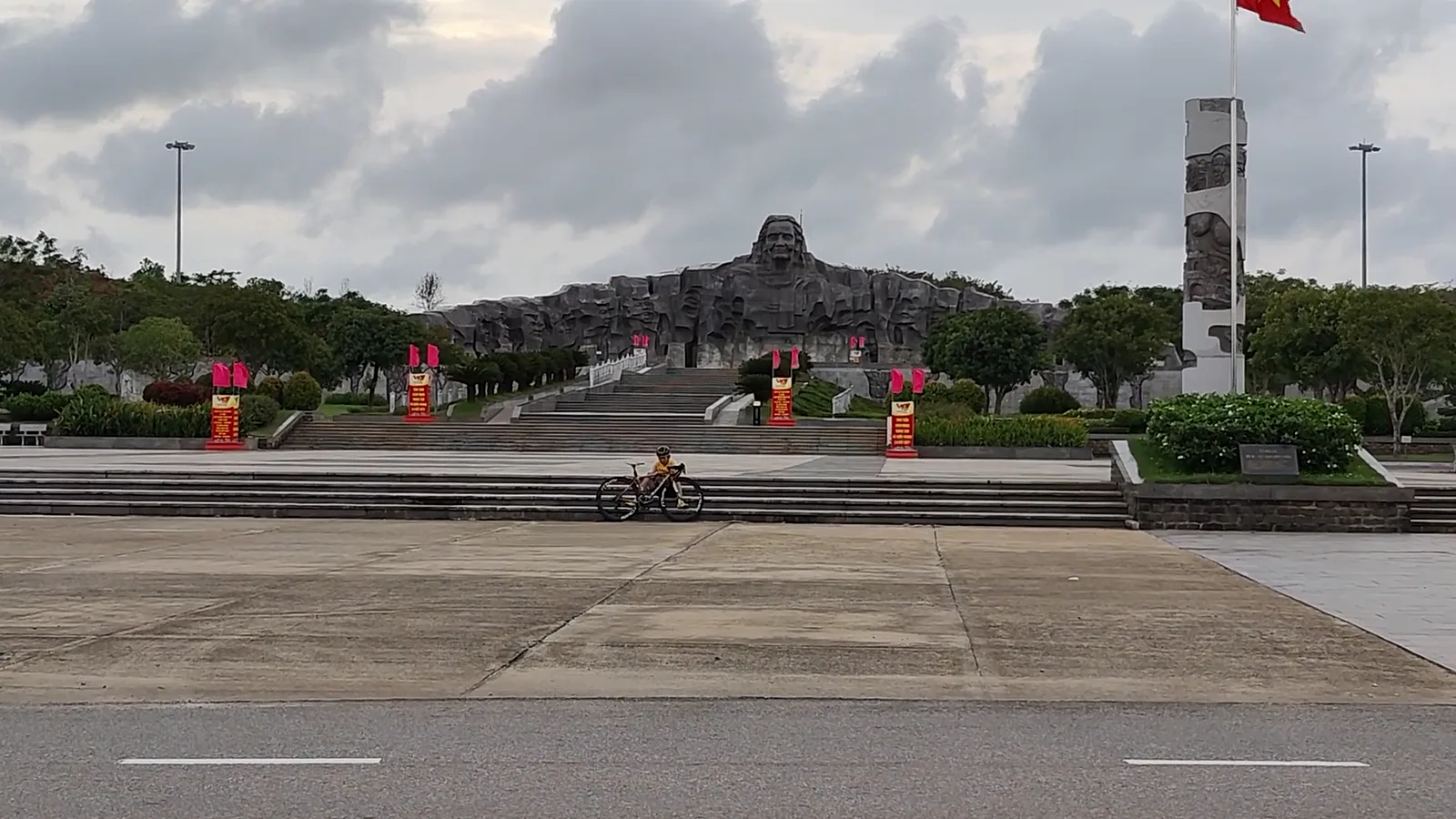Tượng đài Mẹ Việt Nam anh hùng trên núi Cấm Tam Kỳ, khối tượng tạc gương mặt Mẹ giữa quảng trường rộng, một người đi xe đạp ngang qua